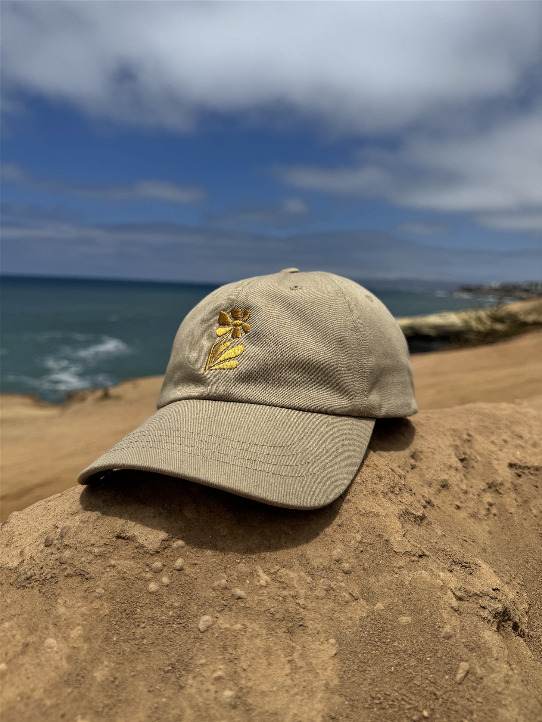Wild Flower Cap khaki dad hat resting on sunlit beach rocks by the ocean