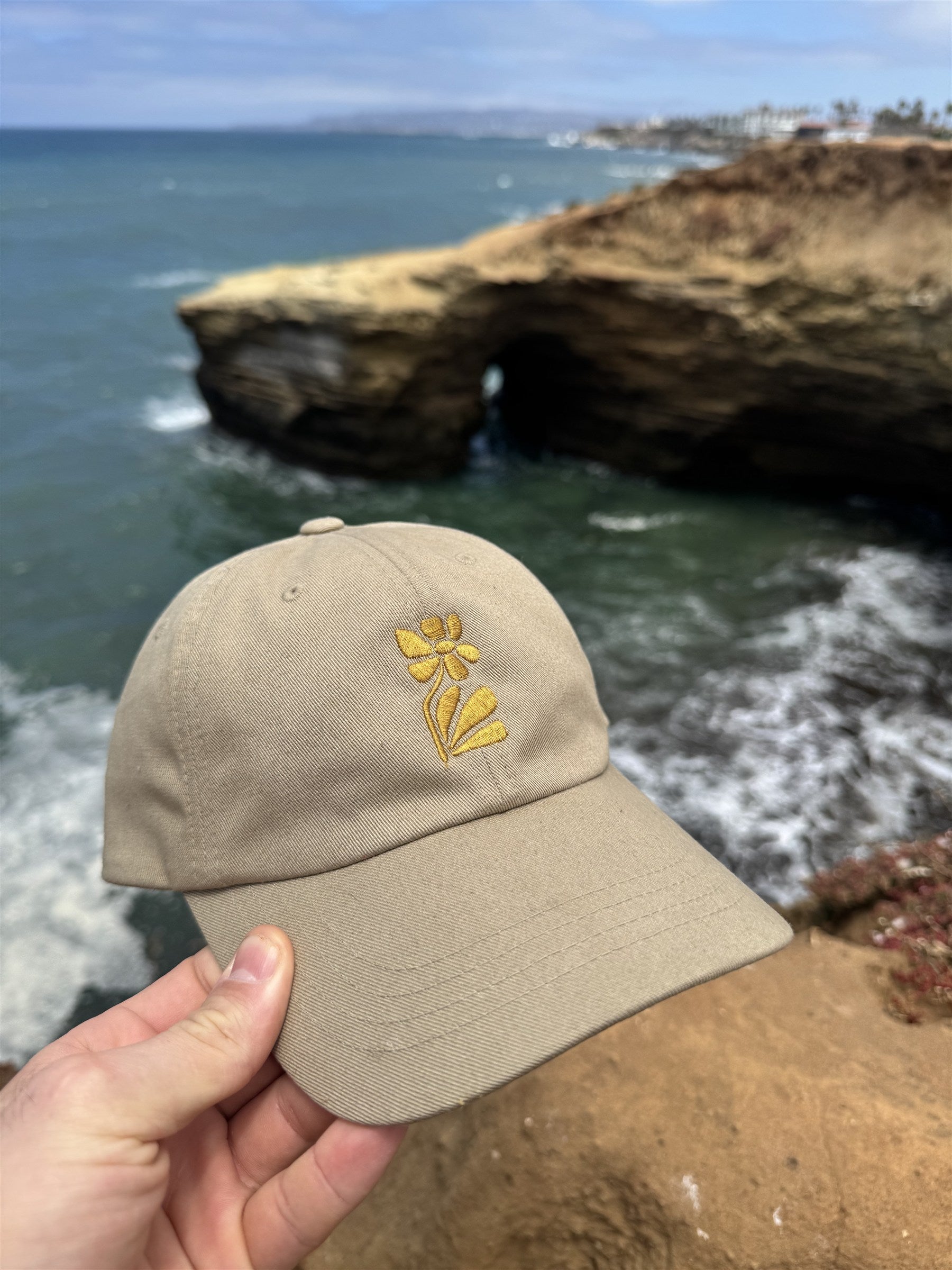 Aura Vita Sola Cap held up over a rocky ocean cove, gold flower embroidery detail